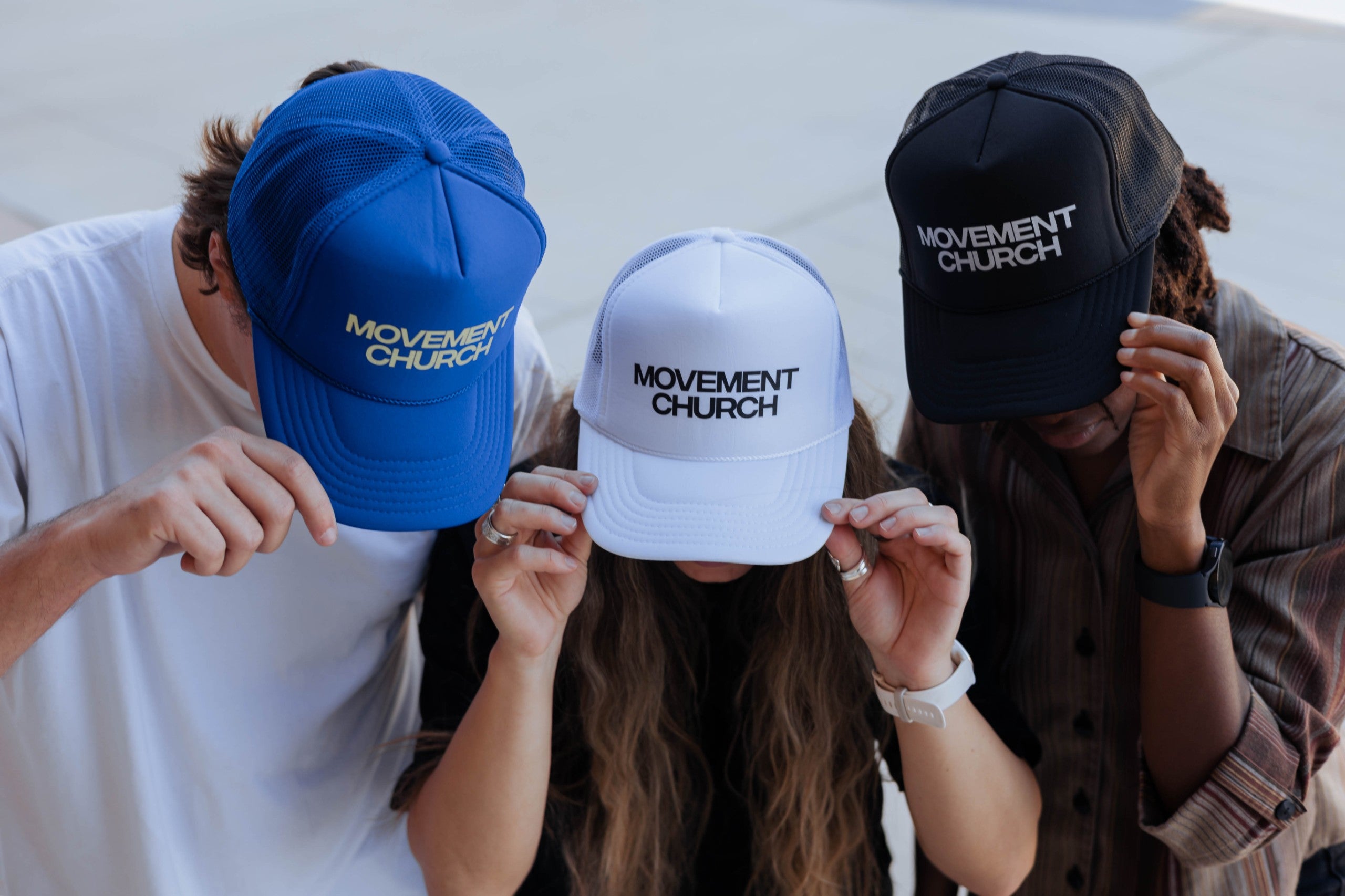 Three people wearing Movement Church trucker hats in blue, white, and black